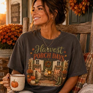 Puede incluir: Una camiseta de manga corta gris carbón con un estampado gráfico de un porche con mecedoras, calabazas y el texto Harvest Porch Days. La camiseta se combina con una taza de cerámica moteada que presenta una pequeña ilustración de calabaza.