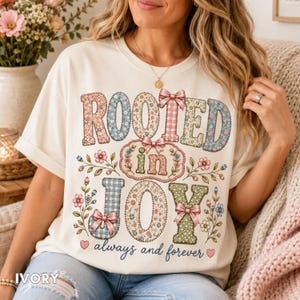 Puede incluir: Una camiseta de color marfil con el texto ROOTED in JOY always and forever. Las letras están rellenas con patrones florales, de cuadros vichy y escoceses en tonos azules, rosas y verdes, acentuadas con pequeños lazos rosas y enredaderas.