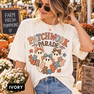 Puede incluir: Una camiseta de manga corta color marfil con un gráfico de tres fantasmas con patrones de patchwork y lazos. El texto dice PATCHWORK PARADE en una fuente negrita multicolor. El diseño incluye pequeñas estrellas y hojas de otoño alrededor.
