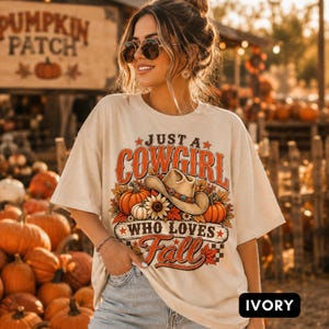 Puede incluir: Una camiseta de color marfil con un estampado gráfico que incluye el texto 'JUST A COWGIRL WHO LOVES Fall'. El diseño presenta un sombrero de vaquero, calabazas, girasoles y hojas de otoño en tonos naranja, marrón y amarillo. Tiene mangas cortas.