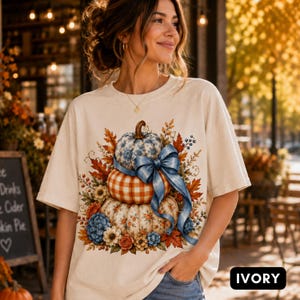 Puede incluir: Una camiseta de manga corta color marfil con un estampado gráfico de tres calabazas apiladas. La calabaza superior tiene un patrón floral azul, la del medio es de cuadros naranjas y blancos, y la inferior tiene un estampado floral. Un gran lazo azul las une.