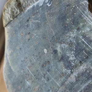 Puede incluir: Primer plano de una roca gris y blanca con una superficie rugosa y texturizada. La roca tiene un brillo plateado y está cubierta de pequeñas manchas oscuras.