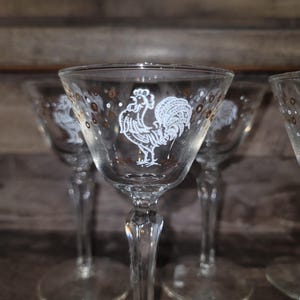 May include: Set of clear stemmed cocktail glasses with a white rooster design. The glasses have a gold rim and gold polka dots. The stems are faceted. The glasses are arranged in a row.