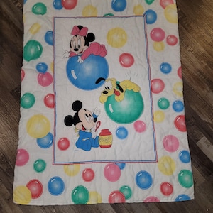 May include: White baby blanket featuring Mickey Mouse, Minnie Mouse, and Pluto. The blanket has a central panel with the characters on blue and green bubbles, surrounded by colorful bubbles. Mickey is blowing bubbles from a jar labeled "Bubbles".