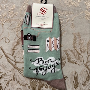 May include: A pair of graphic cotton crew socks with a travel theme. The socks are light green with illustrations of a camera, books, and a map. The words "Bon Voyage" are written in script. The socks are packaged with a black hanger.