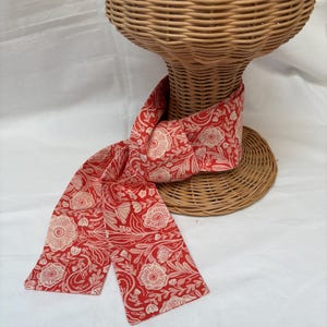 May include: A red scarf with a floral pattern in cream. The scarf is draped over a wicker bust. The floral design features stylized flowers and leaves. The scarf is made of a soft fabric.