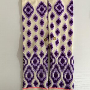 May include: A pair of cream-colored socks with a repeating purple diamond pattern. The socks have a pink stripe at the toe and a gold logo near the ankle. The socks are designed for comfort and style.