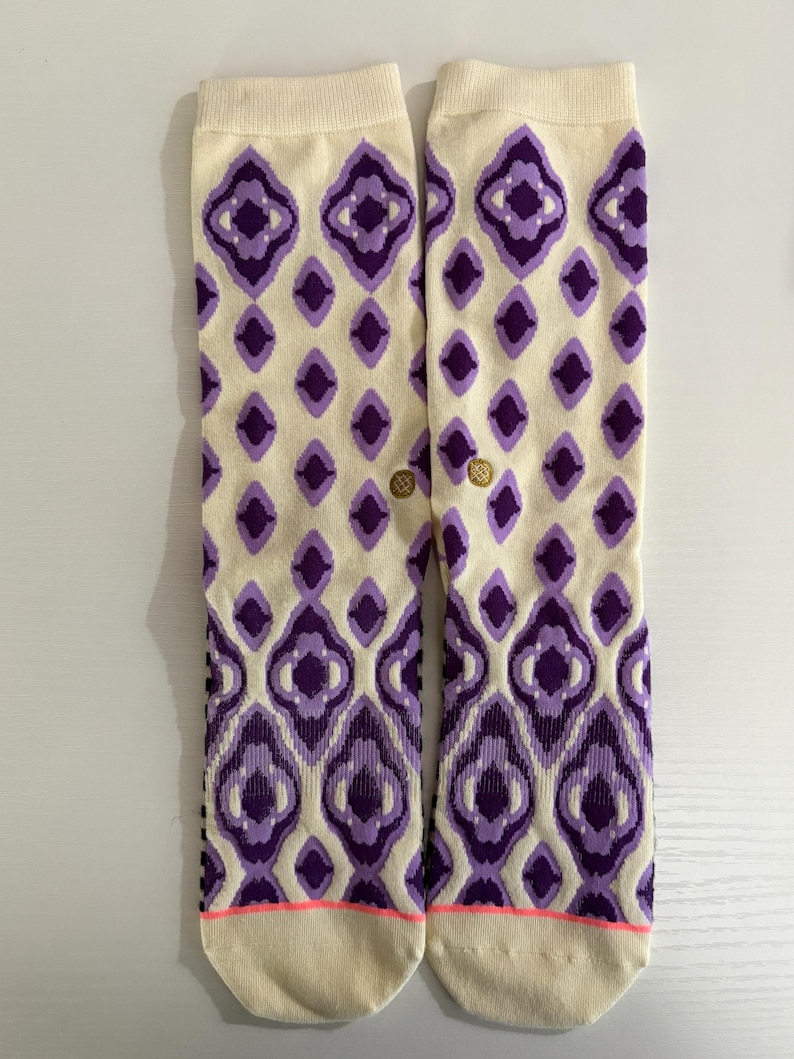 May include: A pair of cream-colored socks with a repeating purple diamond pattern. The socks have a pink stripe at the toe and a gold logo near the ankle. The socks are designed for comfort and style.