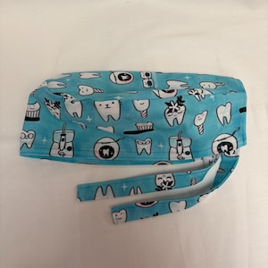 May include: A light blue surgical scrub cap with a pattern of cartoon teeth, toothbrushes, and dental tools. The cap has ties for securing and is designed for medical professionals in dentistry.
