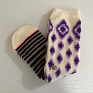 May include: A pair of socks with a cream-colored background. One sock features a pattern of purple diamonds, while the other has black and brown stripes. The toe of the striped sock has a neon pink accent.