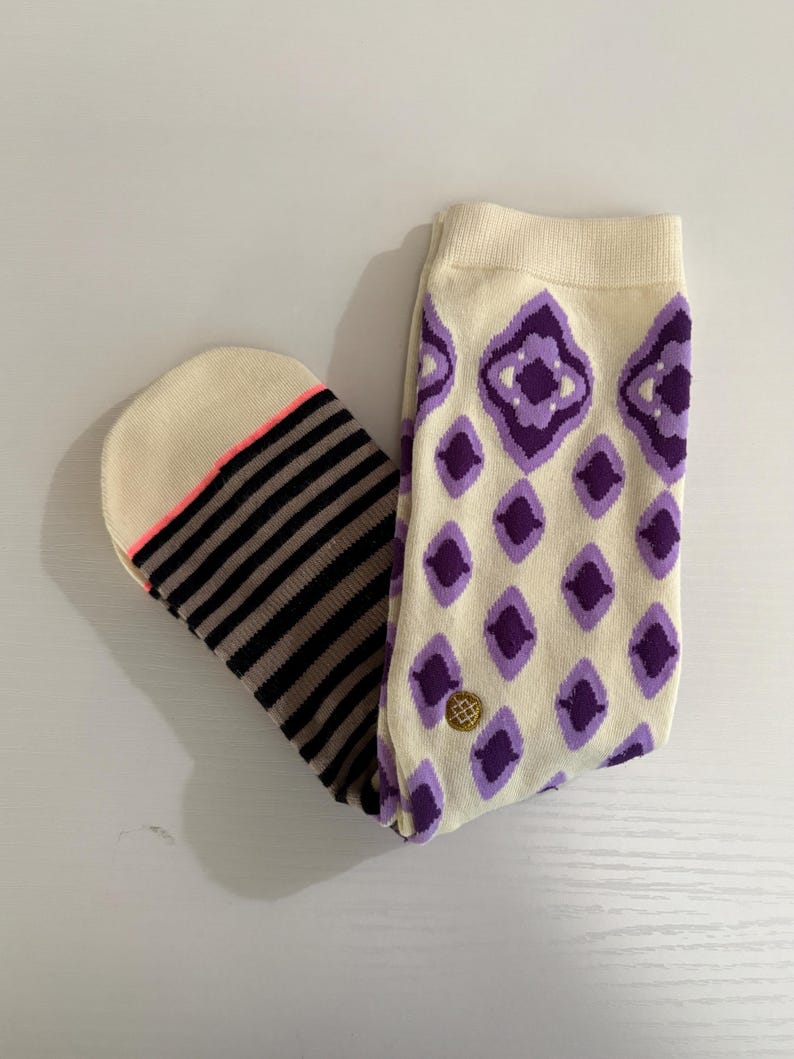 May include: A pair of socks with a cream-colored background. One sock features a pattern of purple diamonds, while the other has black and brown stripes. The toe of the striped sock has a neon pink accent.