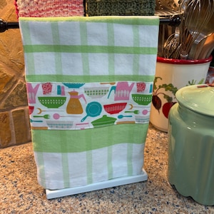 May include: A white kitchen towel with a green and white checkered pattern and a colorful border of kitchenware designs. The towel is hanging on a metal towel rack with two other towels.