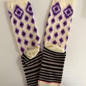 May include: A pair of cream-colored socks with purple diamond patterns on the upper part. The lower part features black and gray horizontal stripes, with a pink accent on the toe.