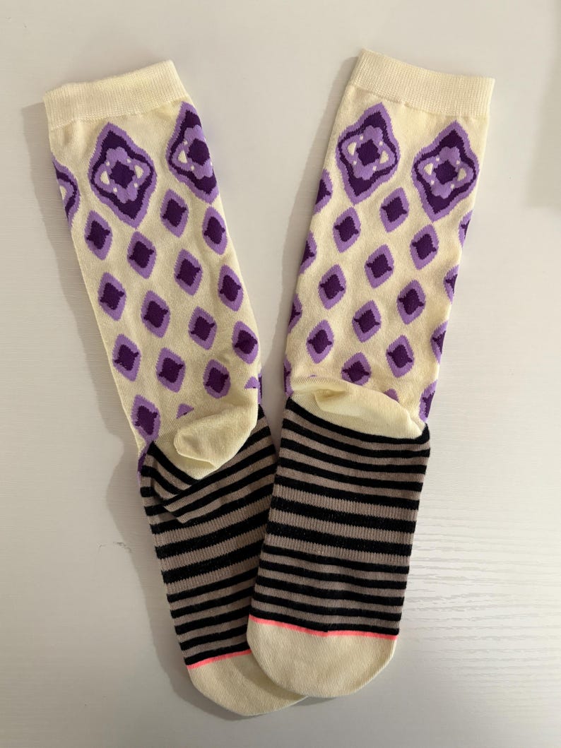 May include: A pair of cream-colored socks with purple diamond patterns on the upper part. The lower part features black and gray horizontal stripes, with a pink accent on the toe.