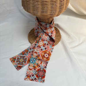 May include: A long, narrow neck scarf featuring a vibrant, repeating geometric pattern in shades of orange, red, teal, and white. The fabric is draped around a woven wicker display stand, showcasing the intricate, symmetrical tile-like design.