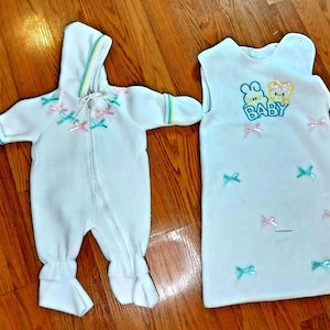 Könnte beinhalten: Ein weißer Fleece-Overall mit Kapuze, Frontreißverschluss, langen Ärmeln und Fußteilen, verziert mit kleinen rosa und türkisfarbenen Schleifen. Daneben liegt ein weißer, ärmelloser Schlafsack mit einem blauen, gestickten BABY-Logo und Schleifen.