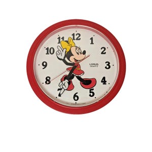 May include: Red-framed Lorus Quartz wall clock featuring Minnie Mouse. The clock face is white with black numerals and hands. Minnie Mouse is depicted in a red dress and yellow bow. The clock measures approximately 10 inches in diameter.