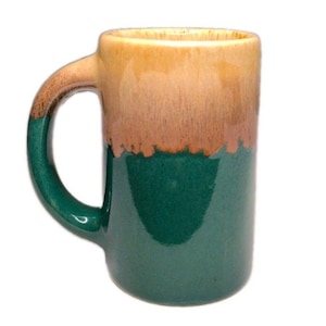 Puede incluir: Una taza de cerámica con un esmalte de dos tonos, que presenta una base de color verde bosque profundo que se funde con una sección superior de color beige arena. La taza tiene un asa curva resistente y un cuerpo cilíndrico con acabado brillante.