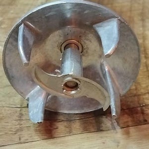 May include: Close-up of a silver immersion blender head with a blade and protective guard. The blender's blade is designed for efficient food processing. The handle is white. The blender is on a wooden surface.