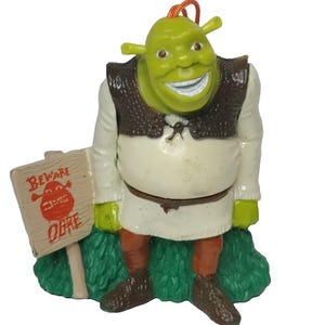 Pode incluir: Uma figura de ogro verde com um rosto sorridente, vestindo uma camisa branca, colete marrom e calças laranja. A figura segura uma placa que diz "Beware Ogre". A figura está em pé sobre uma base verde.