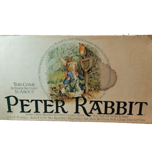 Puede incluir: Caja de juego de mesa vintage de Peter Rabbit. La caja presenta una ilustración de Peter Rabbit con una chaqueta azul, un pájaro y una pala. El texto incluye "Peter Rabbit" y "Based Upon Beatrix Potter's Classic Book".
