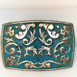Puede incluir: Hebilla de cinturón rectangular plateada con fondo de esmalte turquesa. La hebilla presenta diseños florales y de volutas plateados ornamentados. Los bordes de la hebilla están ligeramente deslustrados, dándole un aspecto vintage.