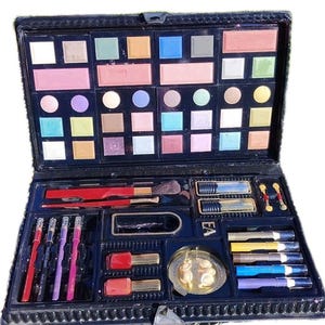 Op de afbeelding: Een zwarte make-up set met een groot palet aan veelkleurige oogschaduw en blush in vierkante en ronde vakjes. Het onderste gedeelte bevat rode en roze nagellakflesjes, diverse make-up kwasten, lippenpotloden en lippenstiften.