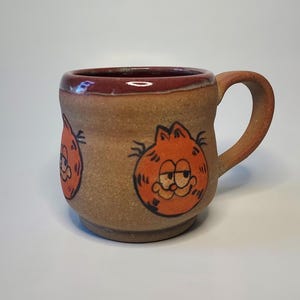 May include: A brown ceramic mug with a red rim and a cartoon image of Garfield the cat. The mug has a handle and is decorated with multiple images of Garfield.