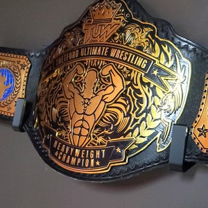 May include: A black leather wrestling championship belt with gold accents. The belt features a large, gold-plated centre plate with a muscular figure, the words "Thetford Ultimate Wrestling", and "Heavyweight Champion". The side plates feature a gold globe with a blue centre and the letters "T.U.W."