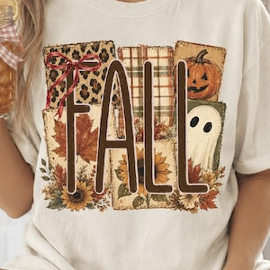 May include: A cream-coloured short-sleeve t-shirt featuring a large graphic with the word FALL in brown letters. The design includes square patches with leopard print, plaid, a pumpkin, a ghost, sunflowers, and autumn leaves with a red bow.