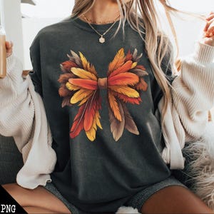 May include: A dark gray t-shirt with a large, colorful bow made of feathers. The bow is made of orange, yellow, and brown feathers. The shirt is a relaxed fit and has a crew neck.