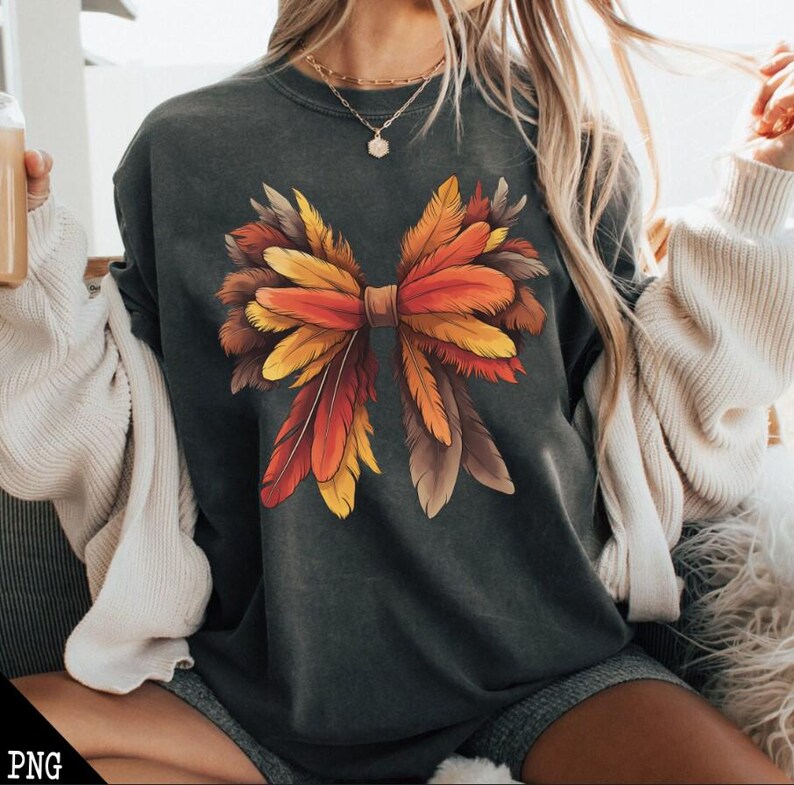 May include: A dark gray t-shirt with a large, colorful bow made of feathers. The bow is made of orange, yellow, and brown feathers. The shirt is a relaxed fit and has a crew neck.