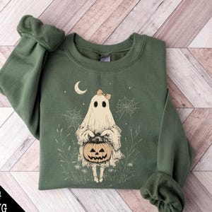 May include: A green sweatshirt featuring a white ghost holding a jack-o'-lantern. The ghost is wearing a pink bow and is surrounded by white spiderwebs and green foliage.