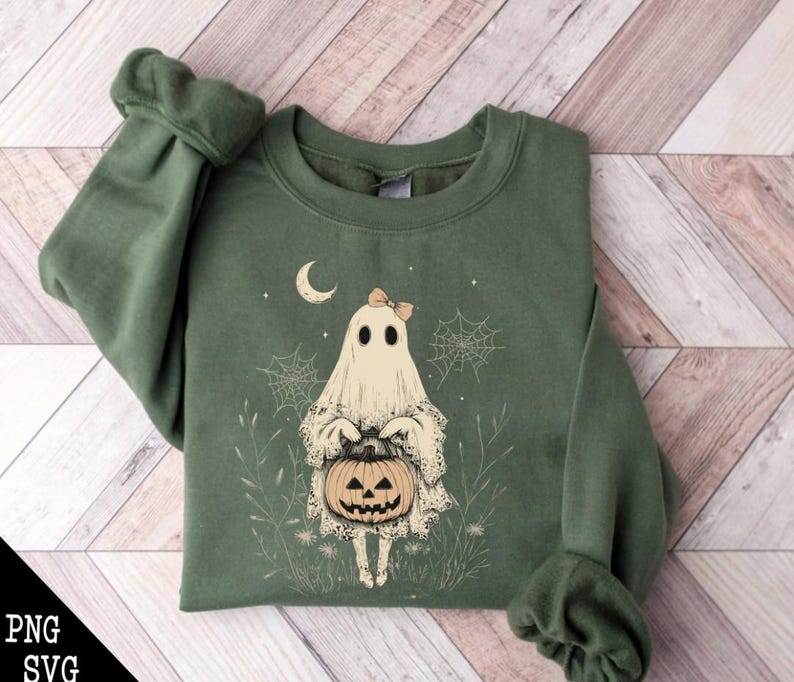 May include: A green sweatshirt featuring a white ghost holding a jack-o'-lantern. The ghost is wearing a pink bow and is surrounded by white spiderwebs and green foliage.