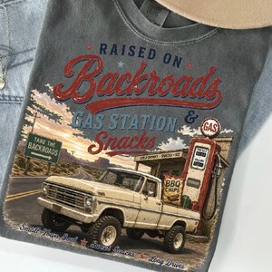 May include: Gray t-shirt with a vintage graphic featuring a cream-colored pickup truck at a gas station. The text reads "RAISED ON Backroads GAS STATION & Snacks". Additional text includes "Take the Backroads", "Small Town Soul", and "Sweet Snacks".