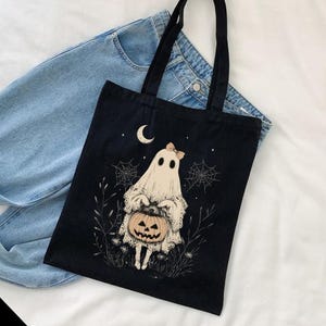May include: Black canvas tote bag featuring a white ghost holding a jack-o'-lantern. The ghost has a pink bow on its head and is surrounded by cobwebs and stars.