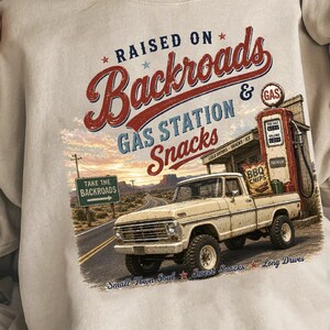 May include: Cream-colored sweatshirt with a graphic featuring a vintage pickup truck, gas station, and the text "RAISED ON Backroads & GAS STATION Snacks." The design includes a road sign that says "TAKE THE BACKROADS."