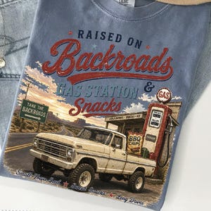 May include: A light blue t-shirt with a graphic featuring a vintage pickup truck at a gas station. The text reads "RAISED ON Backroads GAS STATION & Snacks." Additional text includes "Take the Backroads," "Small Town Soul," and "Sweet Snacks."
