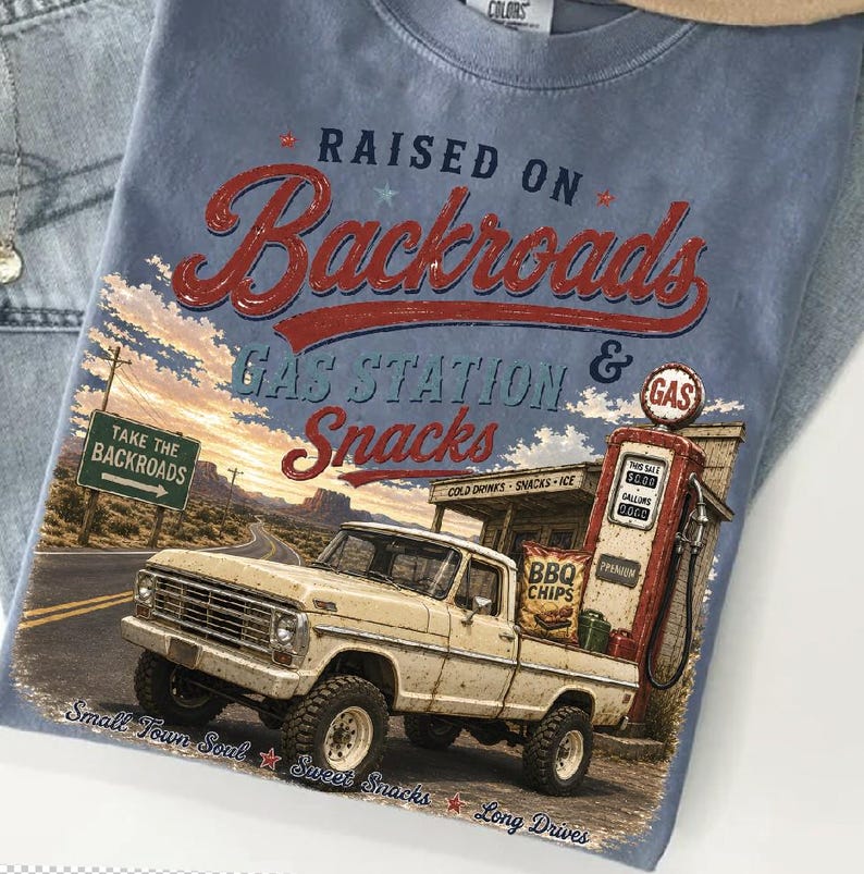 May include: A light blue t-shirt with a graphic featuring a vintage pickup truck at a gas station. The text reads "RAISED ON Backroads GAS STATION & Snacks." Additional text includes "Take the Backroads," "Small Town Soul," and "Sweet Snacks."