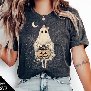 May include: A dark grey t-shirt featuring a white ghost holding a jack-o'-lantern. The ghost has a pink bow on its head and is surrounded by stars and spiderwebs.