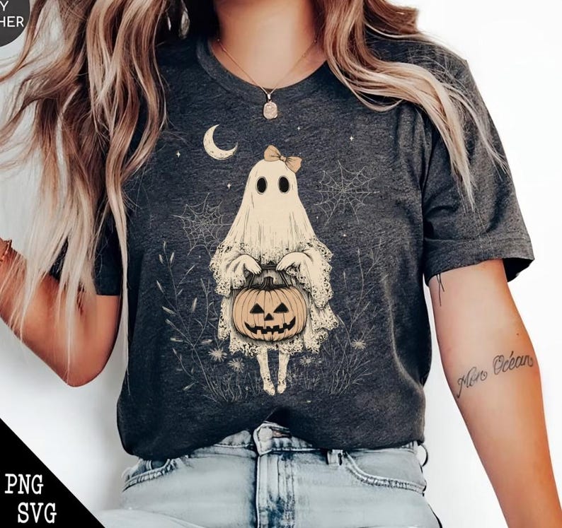 May include: A dark grey t-shirt featuring a white ghost holding a jack-o'-lantern. The ghost has a pink bow on its head and is surrounded by stars and spiderwebs.