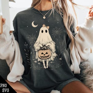 May include: A dark grey t-shirt featuring a white ghost holding a jack-o'-lantern. The ghost is wearing a bow and has a lace trim. The design is surrounded by small white flowers and spiderwebs.