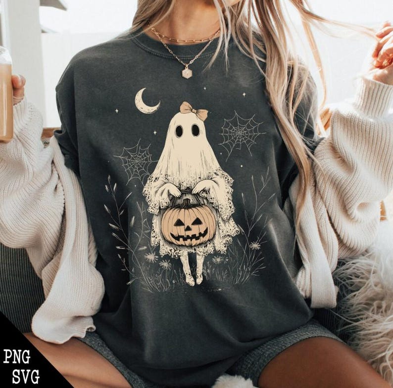 May include: A dark grey t-shirt featuring a white ghost holding a jack-o'-lantern. The ghost is wearing a bow and has a lace trim. The design is surrounded by small white flowers and spiderwebs.