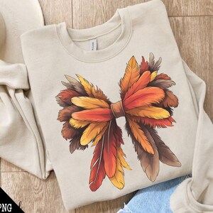 May include: A beige sweatshirt with a large, colorful bow made of feathers. The bow is made of red, orange, yellow, and brown feathers. The feathers are arranged in a way that looks like a bow. The sweatshirt is a great way to show your love for fall.