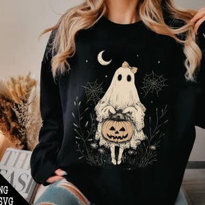 May include: Black sweatshirt featuring a white ghost holding a jack-o'-lantern. The ghost has a pink bow and is surrounded by spiderwebs and stars.