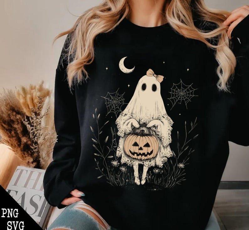 May include: Black sweatshirt featuring a white ghost holding a jack-o'-lantern. The ghost has a pink bow and is surrounded by spiderwebs and stars.