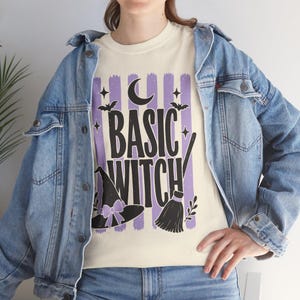 May include: A cream-colored t-shirt featuring the text BASIC WITCH in black, layered over purple vertical brush strokes. The design includes a black witch hat with a purple bow, a crescent moon, small stars, bats, and a broomstick illustration.