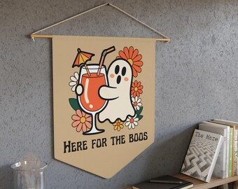 Può includere: Striscione beige con un'illustrazione retrò di un fantasma che tiene in mano un cocktail, circondato da fiori. Il testo "HERE FOR THE BOOS" è stampato sotto. Appeso a un'asta di legno con una corda.