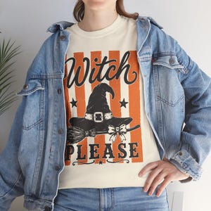 May include: A cream-colored t-shirt featuring a graphic with vertical orange stripes, a black witch hat, a broomstick, and a bow. The text "Witch PLEASE" is printed in black lettering. The shirt is worn under a light blue denim jacket.