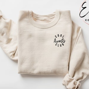 May include: A sand-colored sweatshirt with the text "Cool Aunts Club" in a circle on the front. The sweatshirt is folded and laid flat on a white surface.